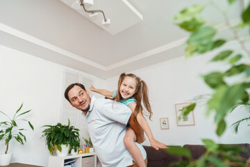 Happy positive cheerful dad is playing with his little charming daughter at home. The man circles and throws the girl up. A dynamic image. Copy space