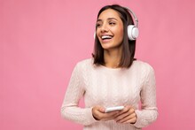 Photo Of Beautiful Joyful Smiling Young Woman Wearing Stylish Casual Clothes Isolated Over Background Wall Holding And Using Mobile Phone Wearing White Bluetooth Headphones Listening To Music And