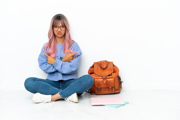 Young student mixed race woman with pink hair sitting one the floor isolated on white background pointing to the laterals having doubts