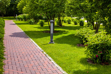 Sticker - straight stone tile pavement in a park landscaped with green grass and bushes with tree bark mulching, garden landscape with ground iron street lights on a sunny summer day.