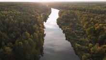 Slow Flight Over Woodland And River At Sunset. The River Surface Reflects The Blue Sky And White Clouds. Beautiful Natural Landscape In Autumn In The Orange Rays Of The Evening Sun.
