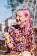 Happy pink-haired teenage hipster girl in a colorful bright T-shirt is eating ice cream on a summer day, with a city fountain in the background.Summer concept.Generation Z style.