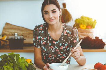 An attractive young dark-haired woman tastes a new recipe for a delicious salad mix while sitting at the table in sunny kitchen