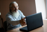 smiling pensive old man with eyeglasses sitting at the desk  and looking at laptop