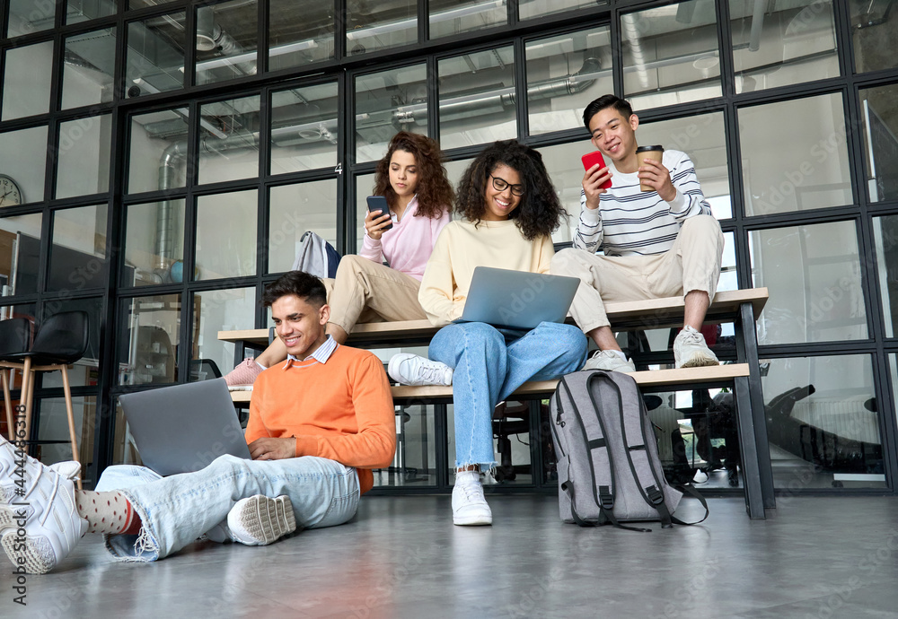 Young happy cheerful multiethnic cool group of high school college students girls and guys sitting at university campus using digital devices social media drinking coffee preparing to seminar classes. - obrazy, fototapety, plakaty 