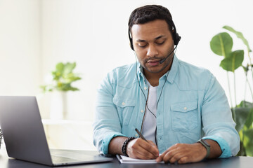 Distance work concept. Young indian man support worker or freelancer uses laptop and headset to communicate with the client, makes notes in a notebook, online learning