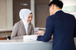 © NVB Stocker - Portrait of muslim woman receptionist at desk in lobby.  Asian business man standing at reception desk and talking to muslim woman receptionist.