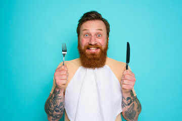 Wall Mural - Happy man with tattoos is ready to eat with cutlery in hand