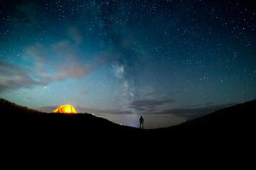 Wall Mural - night photo of a silhouette of a man and an orange tent with light inside, standing on the horizon against the starry sky with the milky way