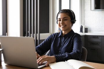 Canvas Print - Happy Indian student taking part in virtual learning webinar from home. Confident employee using headphones and laptop for corporate conference or video call, working at computer, smiling at screen