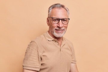 Portrait of handsome bearded senior man smiles pleasantly wears spectacles and casual t shirt looks with self confident expression at camera poses against beige studio background. People and age