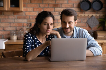 Happy millennial couple using laptop in kitchen, watching show together, making video call, paying bills. Young husband and wife using online app on computer at home, looking at screen, smiling