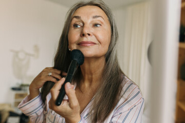 Wall Mural - Smiling elderly woman doing makeup with brush in front of mirror at home. Attractive mature female looking with happiness. Personal care and aging concept