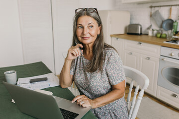 Happy mature female sitting at table and looking away with gladness. Cheerful middle-aged woman working at home. Distance work and remote working concept