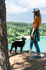 Rear view of young blond woman traveler in cap and ginger t-shirt standing on cliff of blue turquoise lake or river and coniferous forestwith fluffy dog , travel with pets beautiful landscape tourism