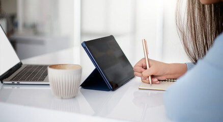 Focus on hand young asian woman holding pen writing on book and learning online with digital tablet and computer laptop