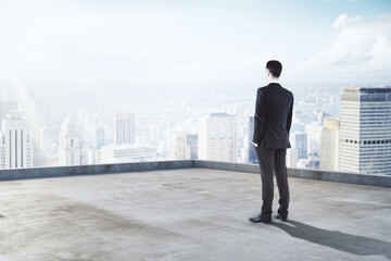 Poster - Pensive young businessman standing on building roof on beautiful sky background with mock up place for your advertisement. Success and future concept.