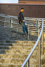 Dressing In Dark Purple Woolen Blazer And Jeans, A Young Guy With Beard And Mustache Is Standing Against A Metal Railing On Stairs, Under Sunshine Of Sunset, Looking At You.