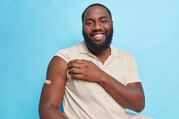 Handsome bearded adult man with dark skin shows arm after vaccination being in good mood feels protected isolated over blue background. Immunization against coronavirus. Health care concept.