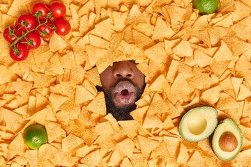 Stunned surprised man with only mouth shown keeps lips rounded makes surprised face buried in crispy mexican nachos chips with red tomatoes green lime and avocado. Unhealthy snacks junk food