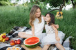 © sofiko14 - Two pretty sisters in casual clothes sitting in embrace on checkered blanket at green garden. Picnic with tasty food and drinks. Relaxation during summertime.