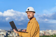 © boytsov - Attractive caucasian bearded engineer using laptop at construction site wearing orange jacket and hard hat as protective workwear. Sunny day with blue cloudy sky and urban skyline. High quality photo