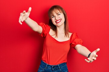 Redhead young woman wearing casual red t shirt looking at the camera smiling with open arms for hug. cheerful expression embracing happiness.