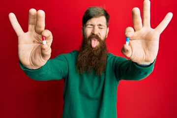 Poster - Redhead man with long beard holding two different pills sticking tongue out happy with funny expression.