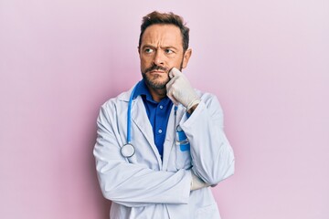 Middle age man wearing doctor uniform and stethoscope serious face thinking about question with hand on chin, thoughtful about confusing idea