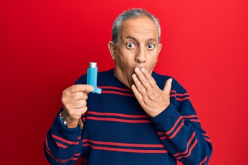 Canvas Print - Senior hispanic man holding medical asthma inhaler covering mouth with hand, shocked and afraid for mistake. surprised expression