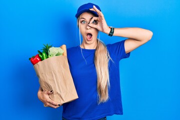 Poster - Young caucasian woman wearing courier uniform with groceries from supermarket doing ok gesture shocked with surprised face, eye looking through fingers. unbelieving expression.