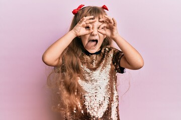 Wall Mural - Little caucasian girl kid wearing festive sequins dress doing ok gesture like binoculars sticking tongue out, eyes looking through fingers. crazy expression.