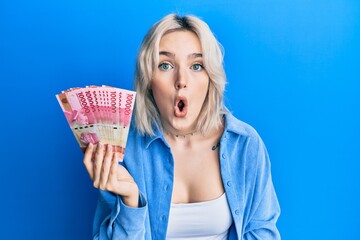 Poster - Young blonde girl holding indonesian rupiah banknotes scared and amazed with open mouth for surprise, disbelief face