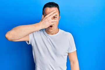 Handsome young man wearing casual white t shirt peeking in shock covering face and eyes with hand, looking through fingers afraid