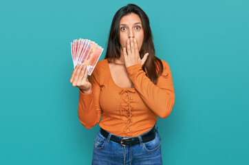 Poster - Beautiful hispanic woman holding norwegian krone banknotes covering mouth with hand, shocked and afraid for mistake. surprised expression