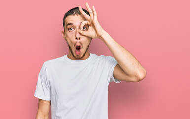 Hispanic young man wearing casual white t shirt doing ok gesture shocked with surprised face, eye looking through fingers. unbelieving expression.