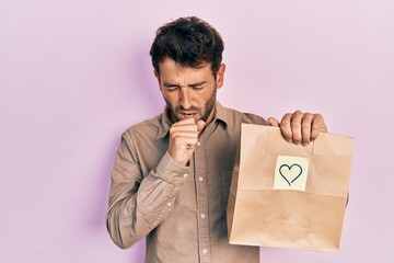 Canvas Print - Handsome man with beard holding delivery paper bag with heart reminder feeling unwell and coughing as symptom for cold or bronchitis. health care concept.