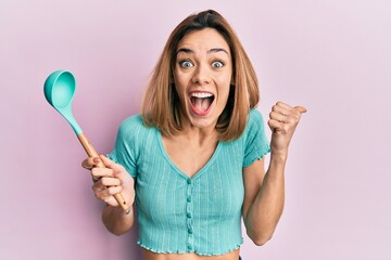 Wall Mural - Young caucasian blonde woman holding silicone soup spoon pointing thumb up to the side smiling happy with open mouth