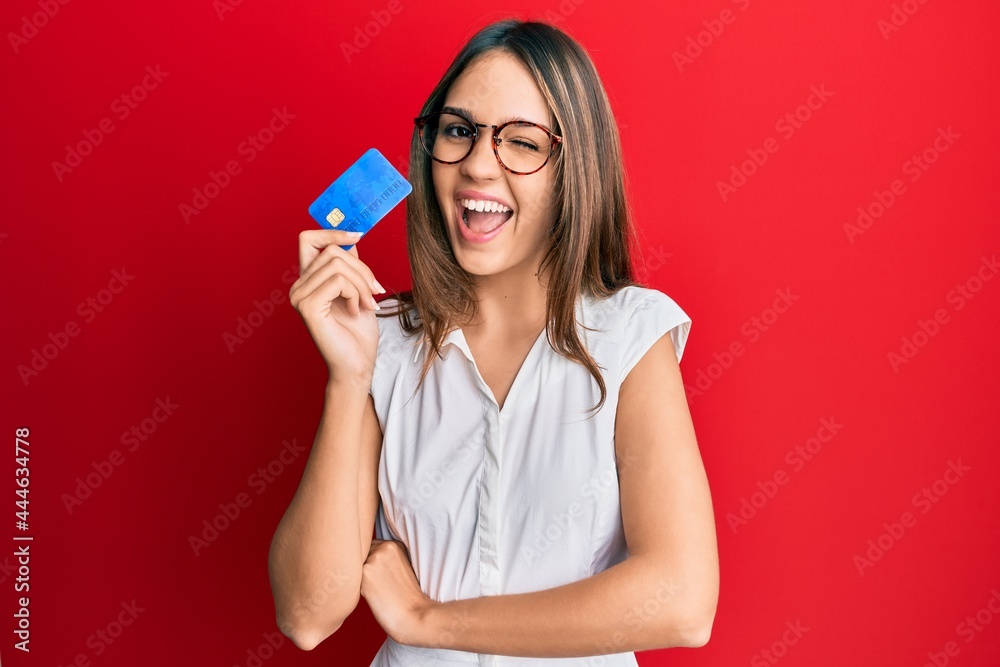 Young brunette woman holding credit card winking looking at the camera with sexy expression, cheerful and happy face. - obrazy, fototapety, plakaty 