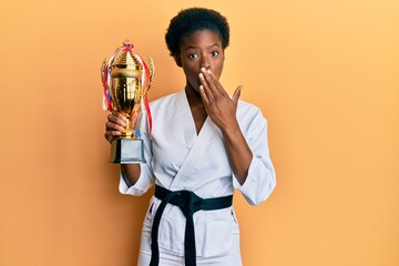 Wall Mural - Young african american girl wearing karate kimono holding trophy covering mouth with hand, shocked and afraid for mistake. surprised expression