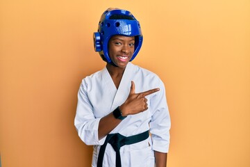 Poster - Young african american girl wearing taekwondo kimono and protection helmet smiling cheerful pointing with hand and finger up to the side