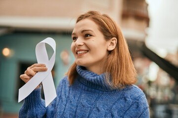 Canvas Print - Young caucasian girl smiling happy holding white awareness ribbon at the city.