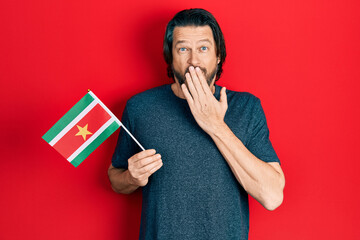 Poster - Middle age caucasian man holding suriname flag covering mouth with hand, shocked and afraid for mistake. surprised expression