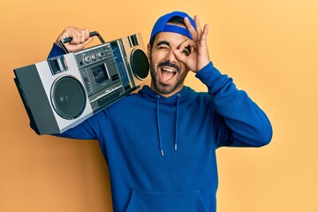 Poster - Young hispanic man holding boombox, listening to music smiling happy doing ok sign with hand on eye looking through fingers