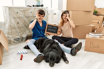 Poster - Young caucasian couple with dog holding our first home blackboard at new house doing ok gesture with hand smiling, eye looking through fingers with happy face.