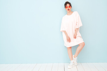 Wall Mural - Portrait of young beautiful smiling female in trendy summer pink clothes. Sexy carefree woman posing near light blue wall in studio. Positive model having fun indoors