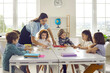 © Studio Romantic - Young female caucasian teacher working with little children group in classroom explaining. Woman tutor and schoolkids joining puzzle. Preschool or primary school education and development
