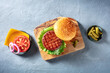 Burger ingredients. Hamburger beef patty, green salad leaf, Cheddar cheese, tomato and red onion, with pickles, overhead shot on a blue background