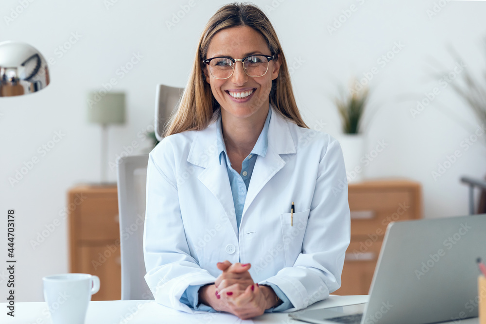 Mature attractive female doctor looking at camera while working sitting in the consultation. - obrazy, fototapety, plakaty 
