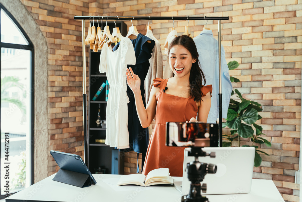 Young and beautiful Asian woman blogger showing clothes in front of smartphone camera while recording vlog video and live streaming at her shop - obrazy, fototapety, plakaty 